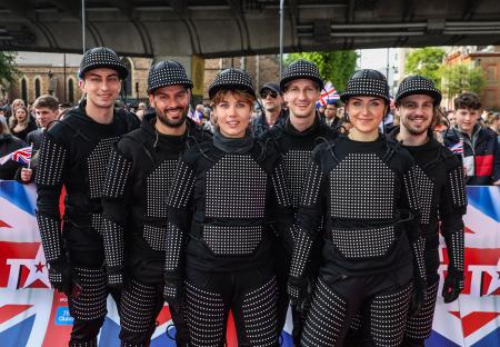 Six performers in black light-up suits on a red carpet.