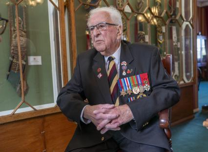 A 100-year-old army veteran wearing medals and sitting in front of a display case.