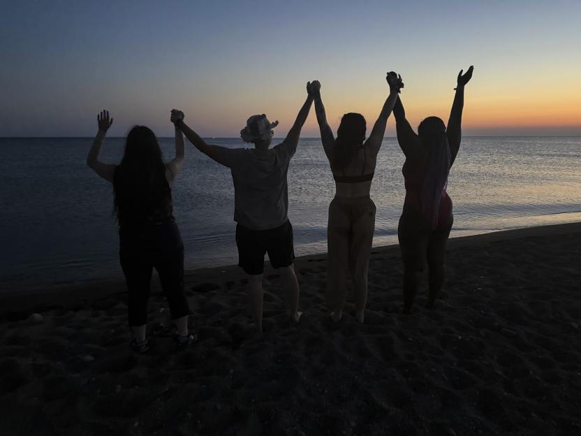 Four people holding hands with arms raised, facing the sea at sunset during a lesbian festival in Lesbos.