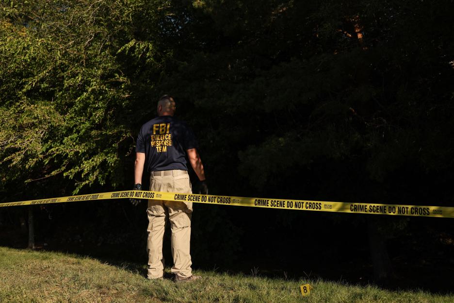 An FBI agent stands behind yellow crime scene tape that reads "CRIME SCENE DO NOT CROSS."