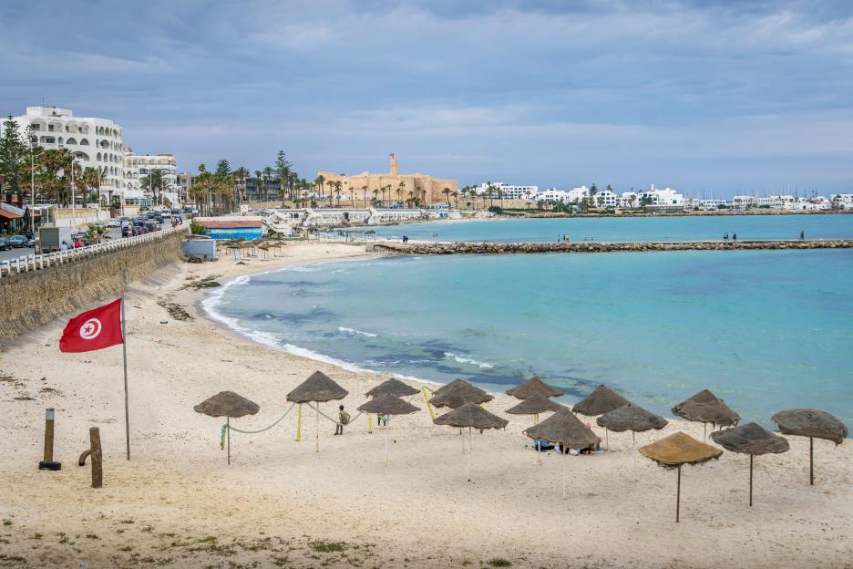 The beach and Mediterranean sea coastline at the resort town of Monastir, Tunisia, with a pier, Tunisian flag, beach umbrellas, and a castle in the background.