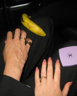 A banana rests on a person's lap, alongside two hands showing wedding rings.