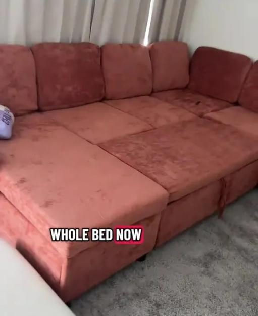 A pink sectional sofa unfolded into a bed with hidden storage, in front of a window with white curtains.