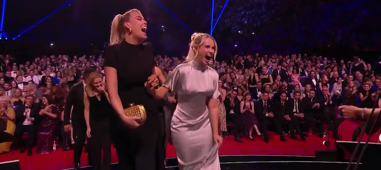 Two women laughing and holding hands while accepting an award in front of a large audience.