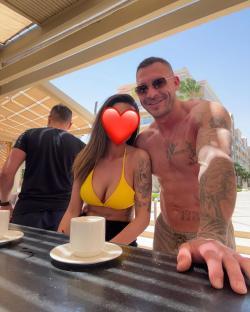 A shirtless tattooed man with sunglasses poses with a woman in a yellow bikini; part of the woman's face is covered by a heart emoji. A man in a black shirt is visible in the background.