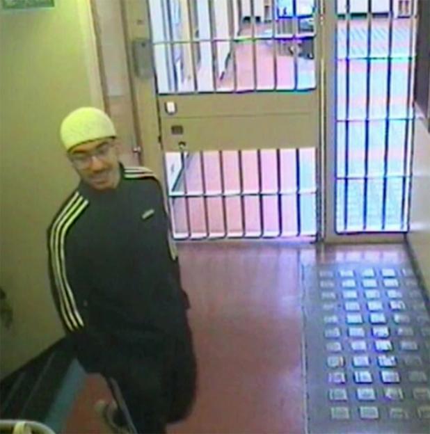 Hashem Abedi, wearing a yellow beanie, glasses, and a black Adidas jacket, smiles in a CCTV image from a prison hallway.