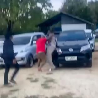 A man in a red shirt fighting with another man near parked cars.