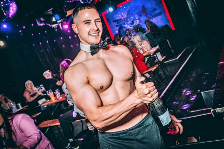 Shirtless male stripper opening a champagne bottle.