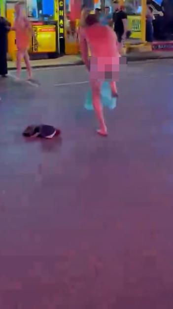 A naked man with pixelated buttocks dances in the middle of a street at night while people watch.