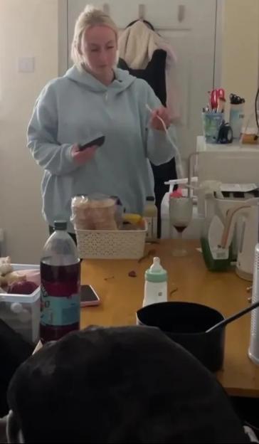A woman in a light blue hoodie holding a phone and a chopstick, with a cluttered table in front of her.