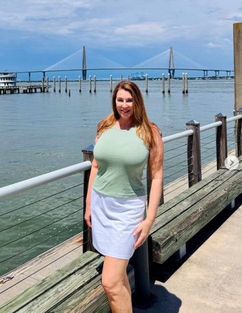 Woman in a sage green top and white skirt poses on a pier with a bridge in the background.