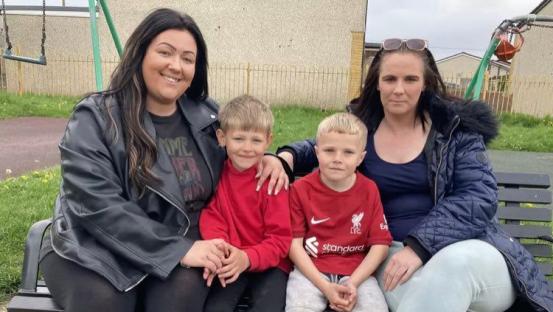 Two young boys and their mothers sitting on a park bench.