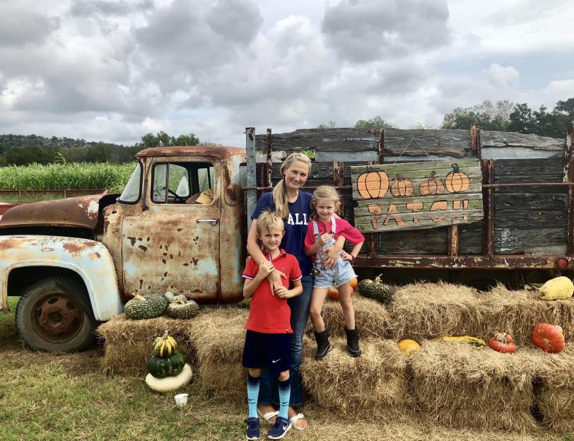 A woman with two children stands in front of a rusty old truck and hay bales decorated with gourds and pumpkins.
