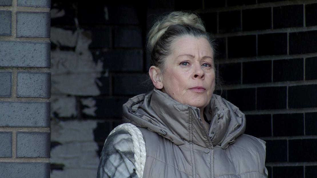 a woman in a grey vest stands in front of a brick wall