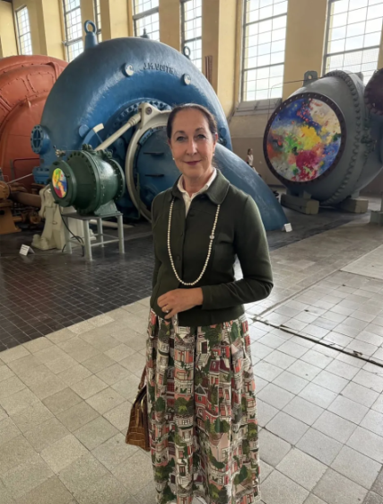 A woman wearing a long, patterned skirt and a dark green jacket with a pearl necklace stands in front of large industrial machinery.