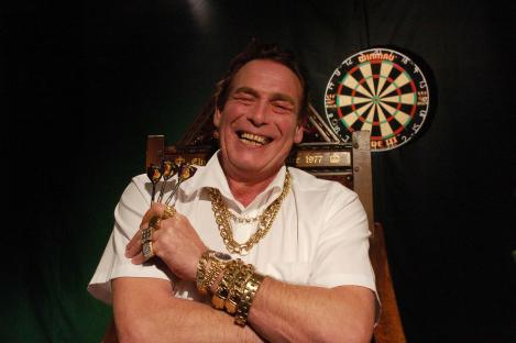 A smiling darts player wearing many gold chains and rings, holding darts. A dartboard is in the background.