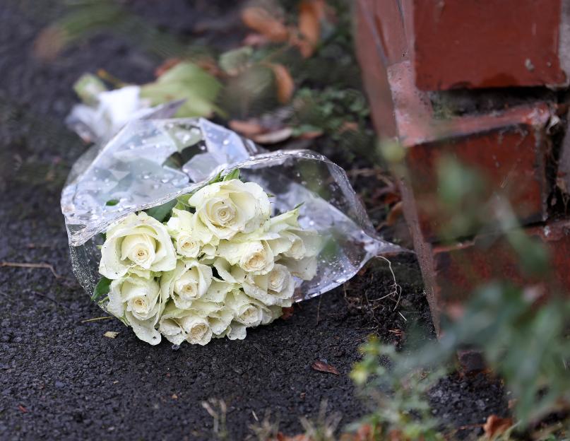 A bouquet of white roses wrapped in clear plastic lies on the asphalt, next to a red brick wall.