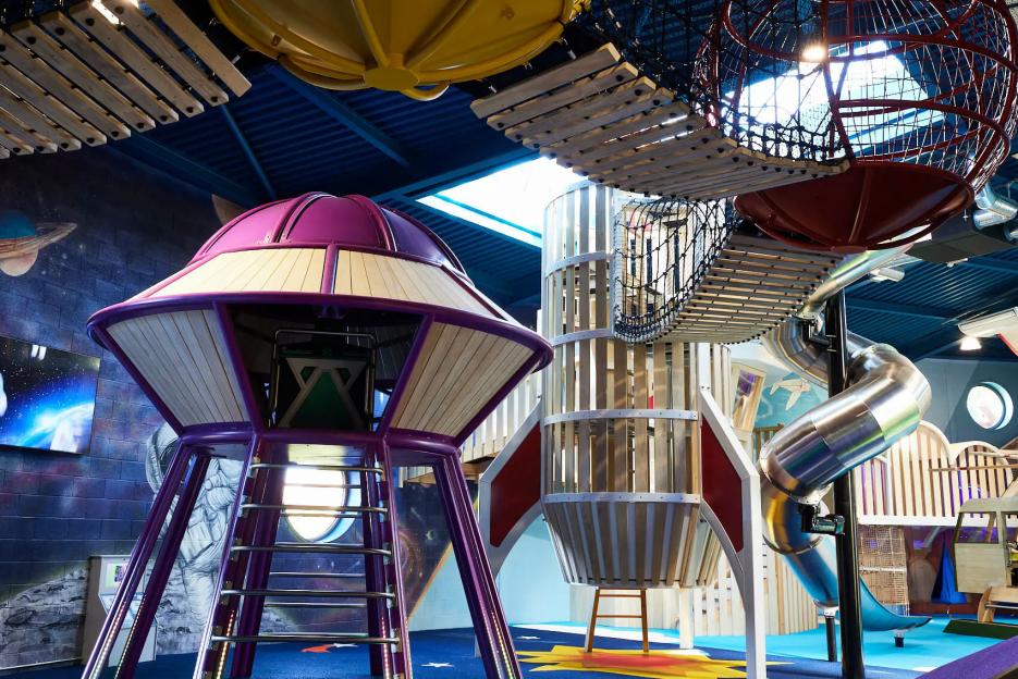 Indoor playground with space-themed structures including a purple UFO climbing frame, a large wooden rocket, and an elevated rope bridge.