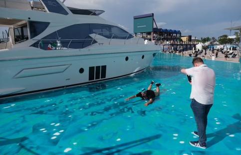 A person lies on a fake water surface next to a yacht while another person takes a photo.