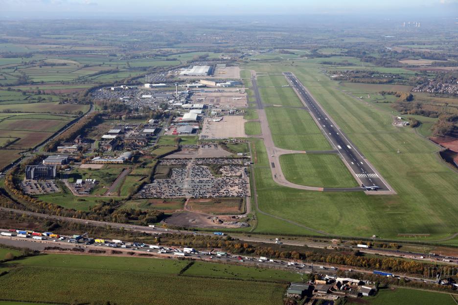 Aerial view of East Midlands Airport in Derby, UK, showing the runway, airport buildings, parking lots, and surrounding countryside with a busy highway.