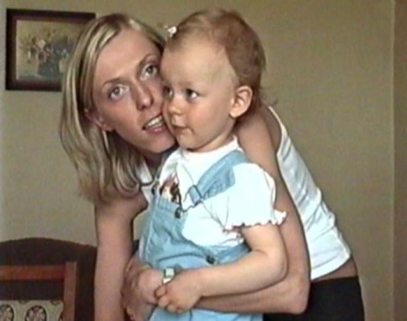 Woman in white tank top holding a baby in blue overalls.