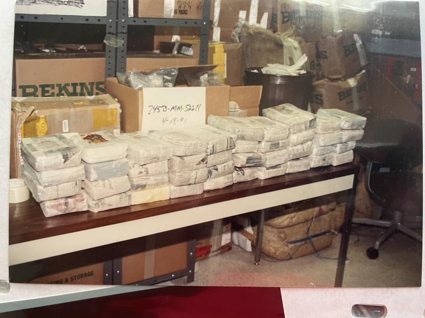 Bundles of drugs wrapped in paper and plastic sitting on a long table in a room full of boxes.