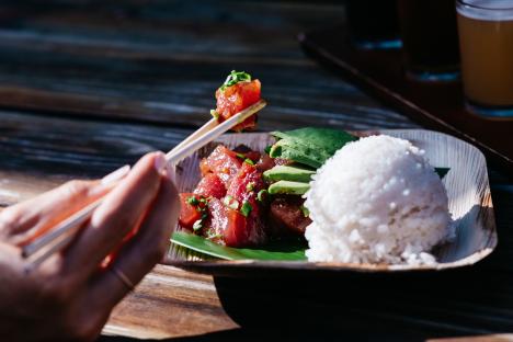 A person uses chopsticks to eat poke, served with rice and avocado.