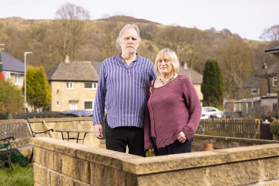 A retired couple stands in their garden, looking at the camera.
