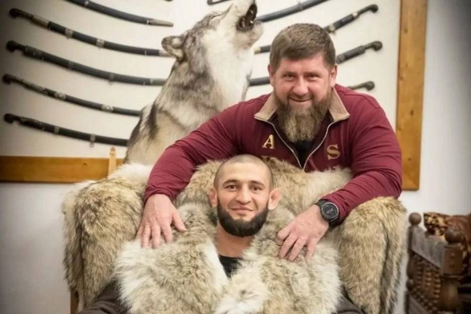 Two men posing for a photo, one wearing a fur coat. A taxidermied wolf is behind them.
