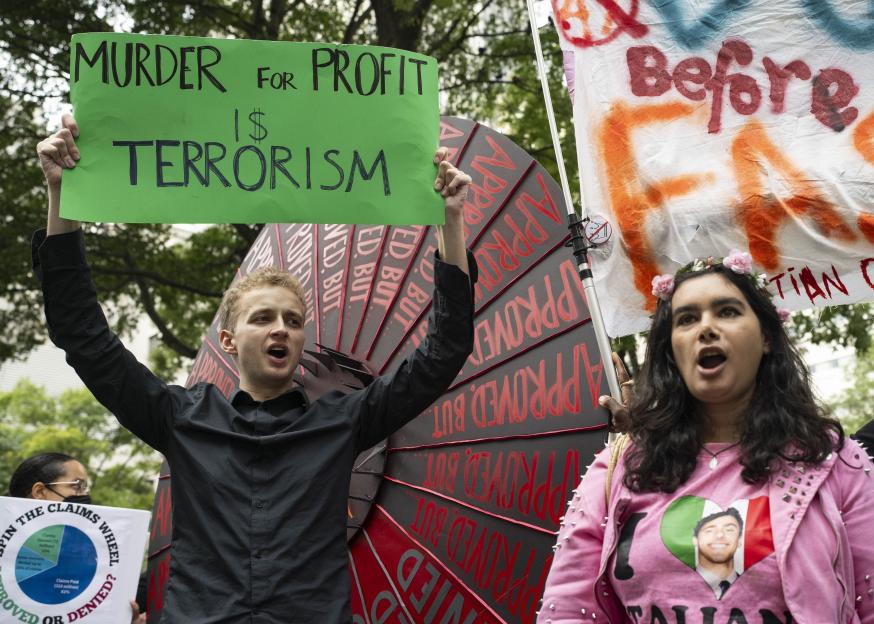 People rally in support of Luigi Mangione, with one holding a sign that reads "MURDER FOR PROFIT IS TERRORISM" and another wearing a shirt that says "I LOVE ITALIAN."