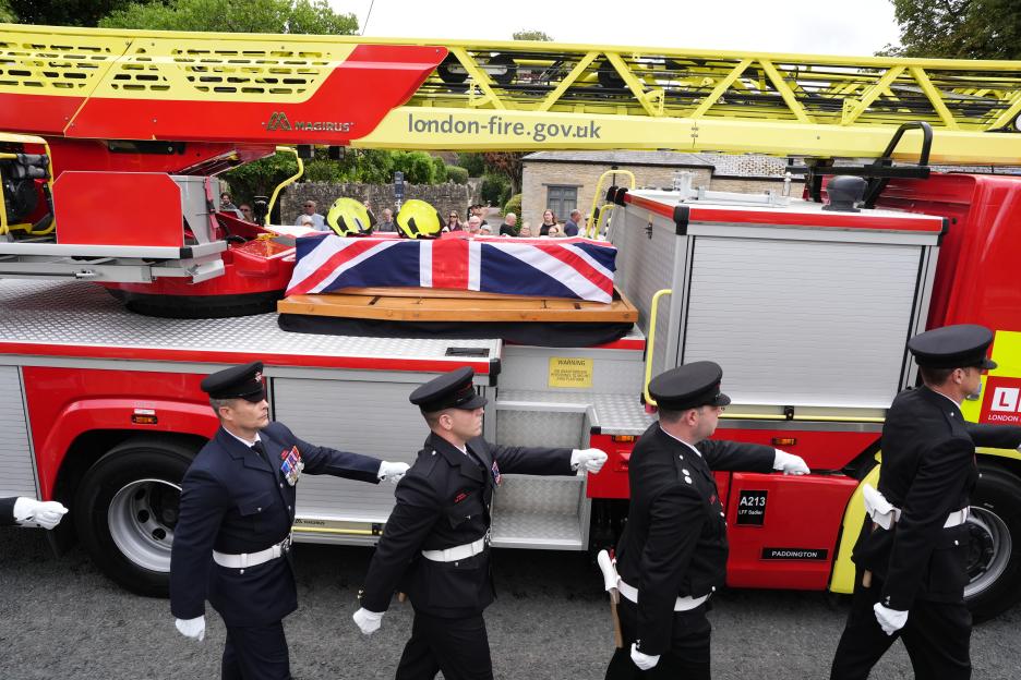Firefighters escorting a coffin draped with a Union Jack on a firetruck.