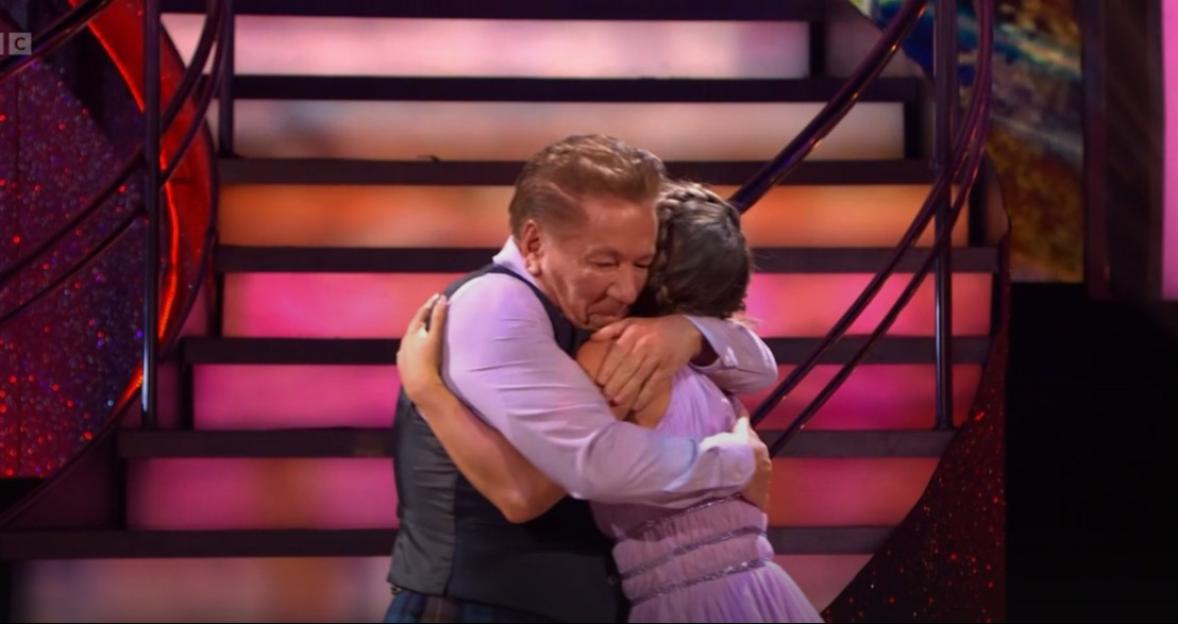 A man and a woman in formal wear hug on a stage with lighted stairs in the background.