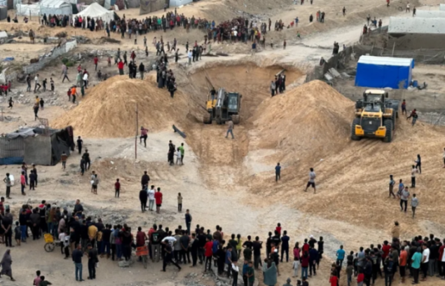 A crowd gathers around excavators digging in a sandy landscape.