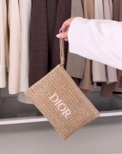 Hand holding a beige Dior raffia clutch bag in front of a closet.