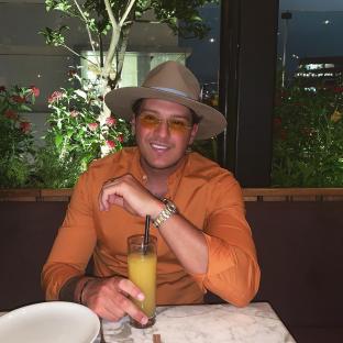 Man in orange shirt and hat sitting at a table with a drink.
