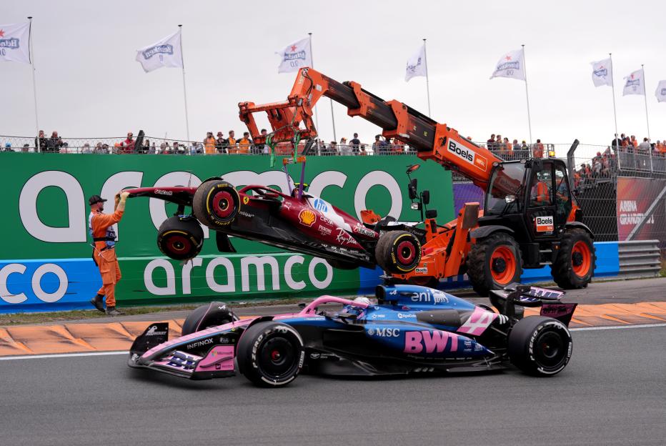 A crane lifts a crashed Formula 1 car from a racetrack as another car drives past.
