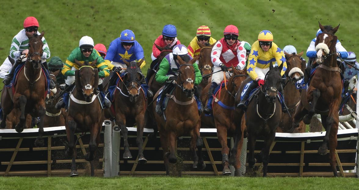 Multiple horses and jockeys jumping over a hurdle during the Coral Cup Handicap Hurdle at Cheltenham Festival.