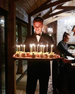 A waiter carrying a tray of cupcakes with lit candles.