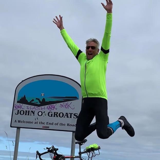 Man in neon green cycling gear jumping in front of John O' Groats sign.