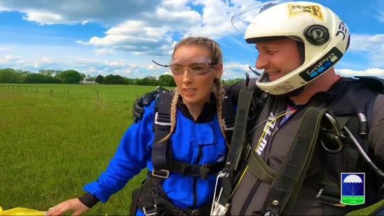 A woman and man in skydiving gear after a jump, in a field.