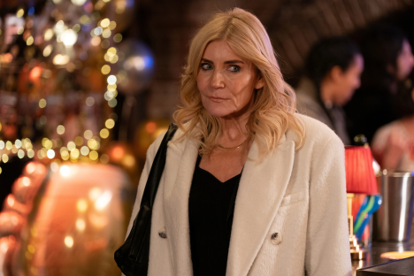A blonde woman in a white coat looks to her left.