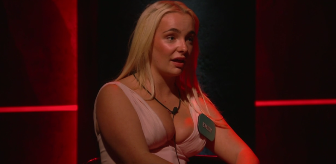 Emily in a light pink top with a microphone on a dark stage with red lighting.