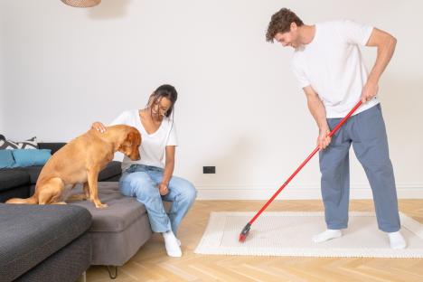 A couple cleans a rug with a broom while their dog sits on a couch.