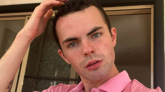 Portrait of a young man with light eyes and dark hair, wearing a pink shirt.