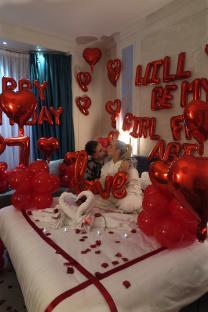 Couple kissing on a bed decorated with red balloons and rose petals.