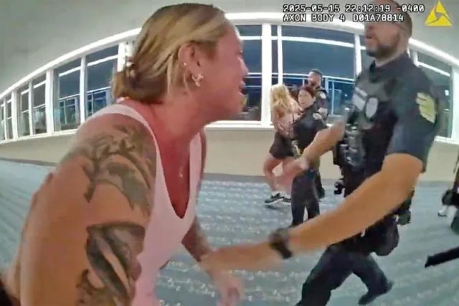 A woman in a pink top with tattoos on her arm, looking distressed, is being approached by police officers in an airport.