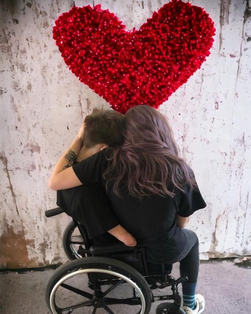 A woman sitting in a wheelchair embraces a man from behind, beneath a large red heart decoration.