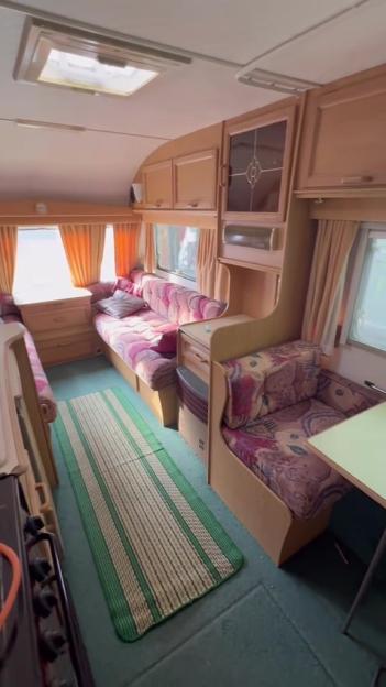 Interior of an old caravan with patterned cushions, orange curtains, and wooden cupboards.