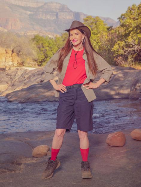 Janice Dickinson from "I'm a Celebrity... South Africa" stands in a tan shirt, red t-shirt, blue shorts, red socks, and hiking boots, with a wide-brimmed hat, against a backdrop of a river and mountains.