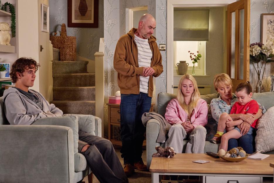 A family sits in their living room, receiving news from a man standing.